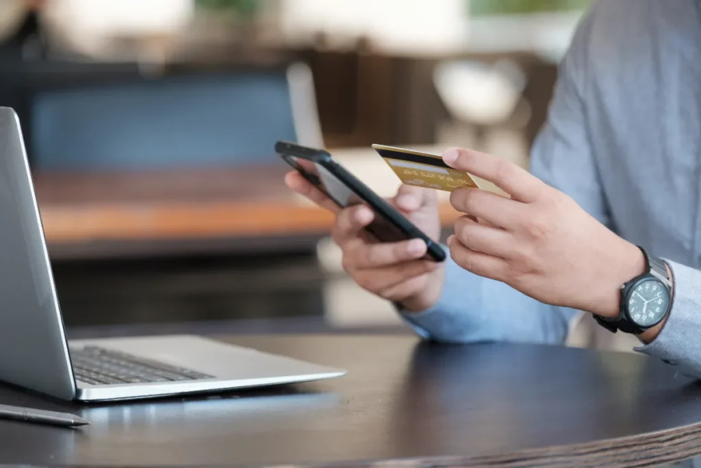 Person hält ein Smartphone und eine Kreditkarte neben einem Laptop